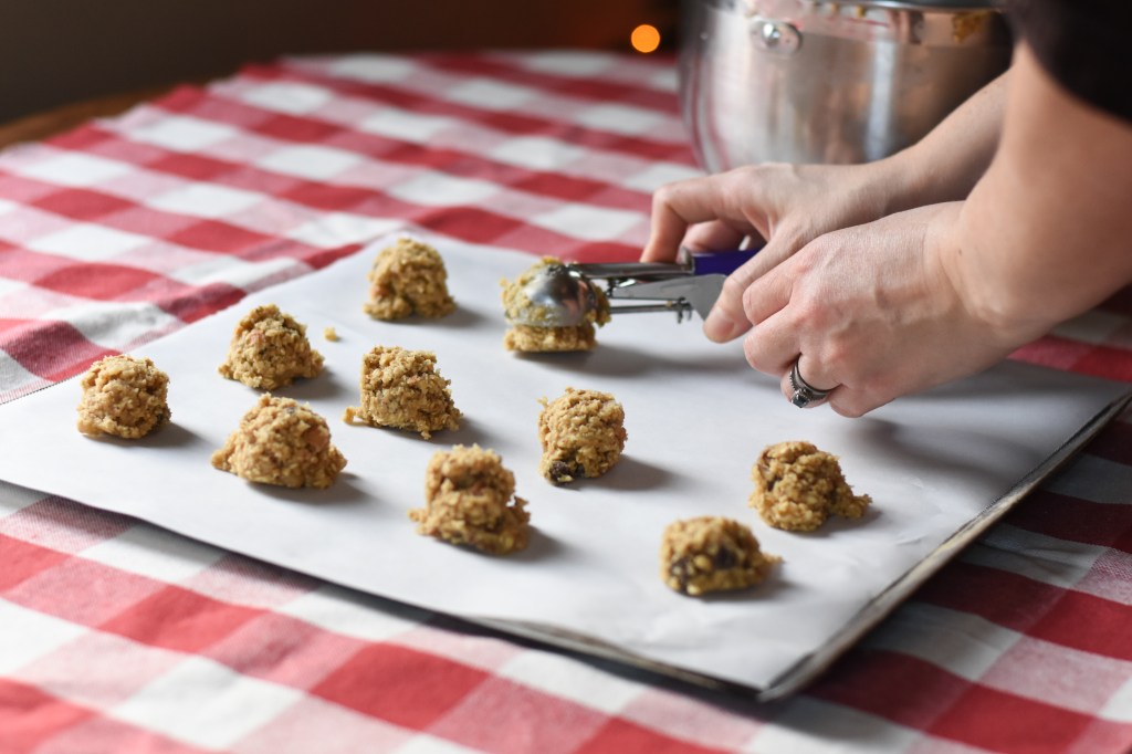 Hands are scooping cookie dough onto cookie sheet. Several scoops are already on the tray.