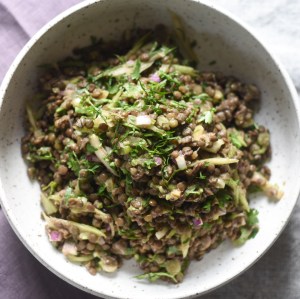 Closeup of green lentil salad with bias cut celery, and lots of fresh herbs. Salad has not meat or dairy and has a purple and gray background.